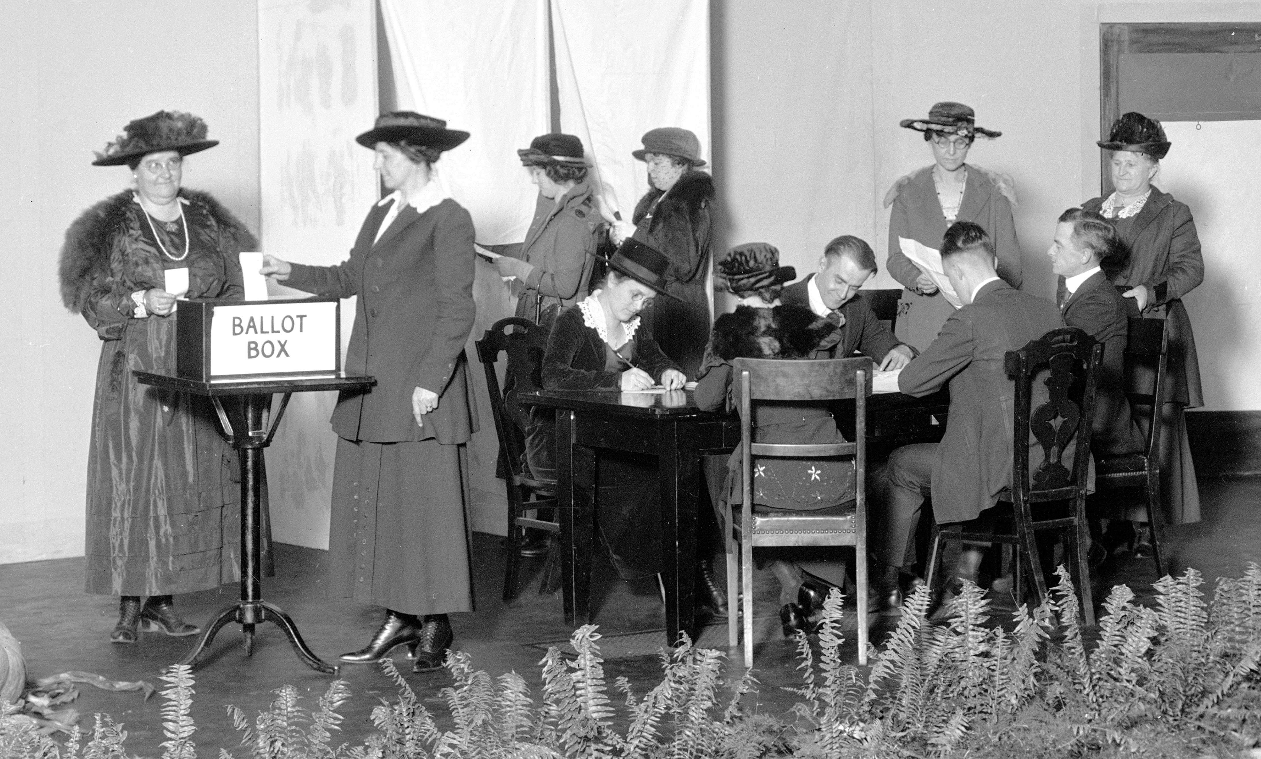 Black and white photo of a table of men and women sitting down and voting on a ballot next to a woman putting a ballot into a ballot box.